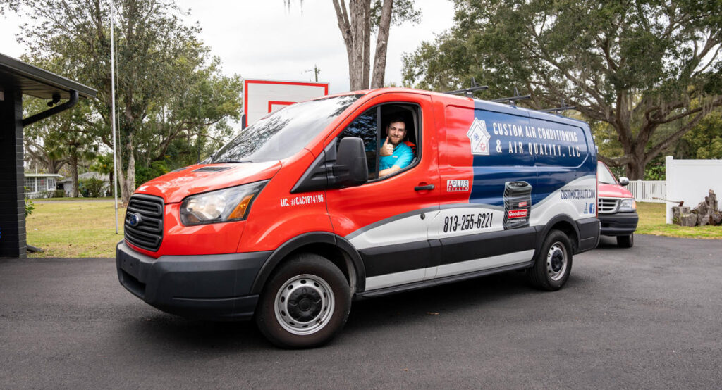 A Custom Air Conditioning & Air Quality technician gives a thumbs up from the driver’s seat of a branded company van showcasing the company's logo contact number and services