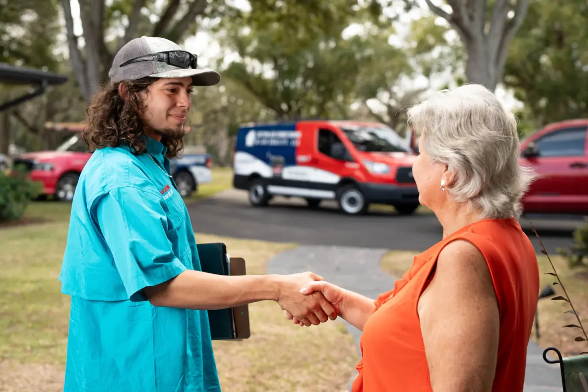 A Custom Air Conditioning & Air Quality technician shakes hands with a customer outside her home, with a branded company van visible in the background