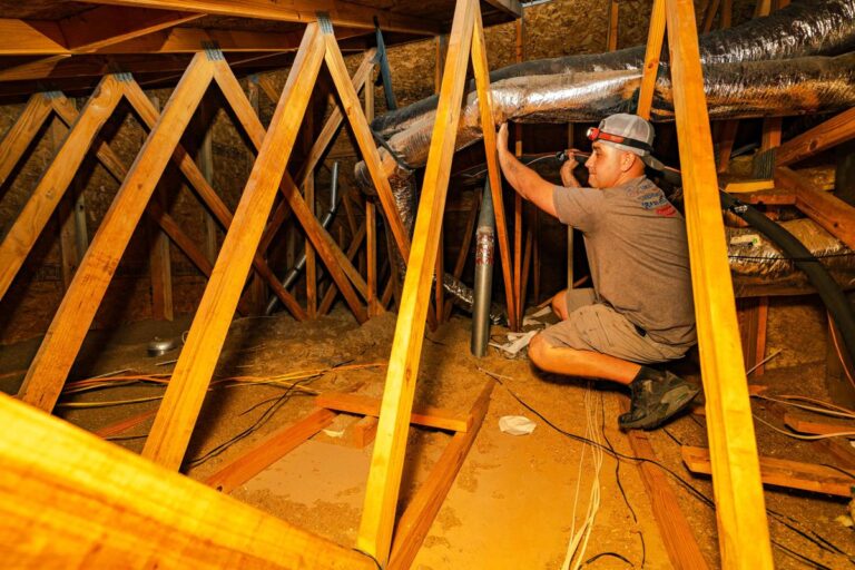 A Custom Air Conditioning & Air Quality technician works in an attic space securing ductwork while wearing a headlamp ensuring proper installation in a tight wood framed area