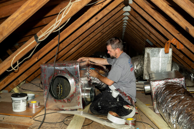 A technician works in a large attic sealing and insulating HVAC ductwork with metallic tape The technician is kneeling next to a large insulated air handler