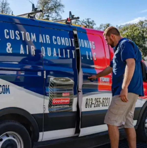 A technician opens the side door of a Custom Air Conditioning & Air Quality van. The van features a colorful design with company branding, including a large 'Goodman' logo and the company’s contact number: 813-255-6221.