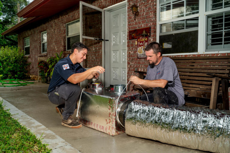 Two Custom Air Conditioning & Air Quality technicians work on sealing an insulated air duct with metallic tape