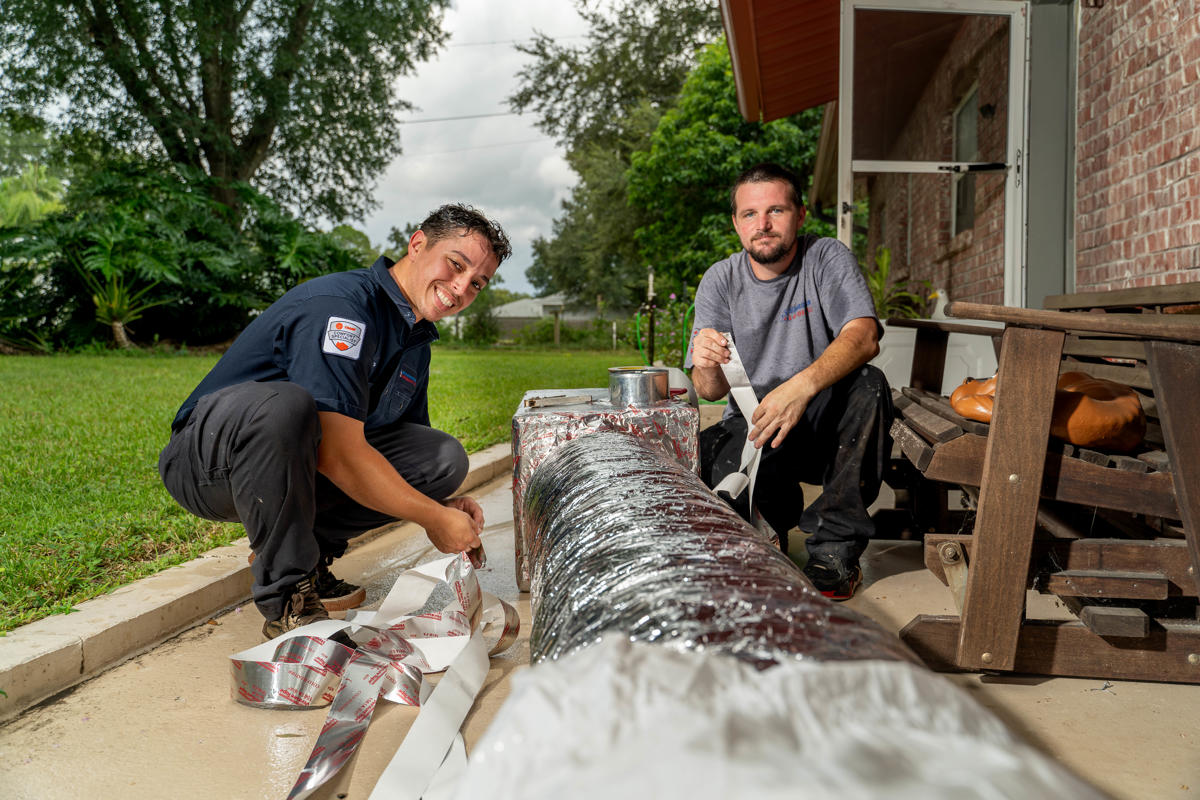 Two Custom Air Conditioning & Air Quality technicians work on wrapping and sealing an insulated air duct outside a home both smiling and focused on the task